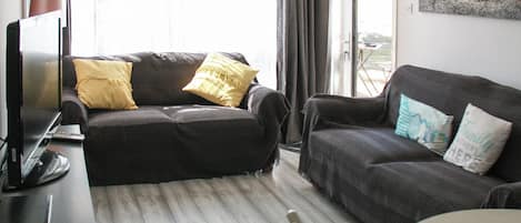 A living room with a dark-colored sofa, a smaller couch, a TV, and a laptop on a desk.