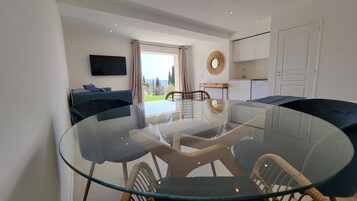 Una sala de estar moderna con una mesa de comedor de vidrio, sillas tapizadas en azul oscuro, un televisor de pantalla plana montado en la pared y vistas a un jardín a través de un ventanal grande.