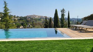 A swimming pool with a wooden deck and lounge chairs, surrounded by greenery and overlooking a hillside town. - Ideal BnB (Saint-Paul-de-Vence)