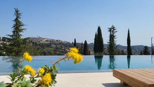 A clear view of a swimming pool with a wooden deck and a bench, overlooking a hillside with cypress trees and a distant town. - Ideal BnB (Saint-Paul-de-Vence)