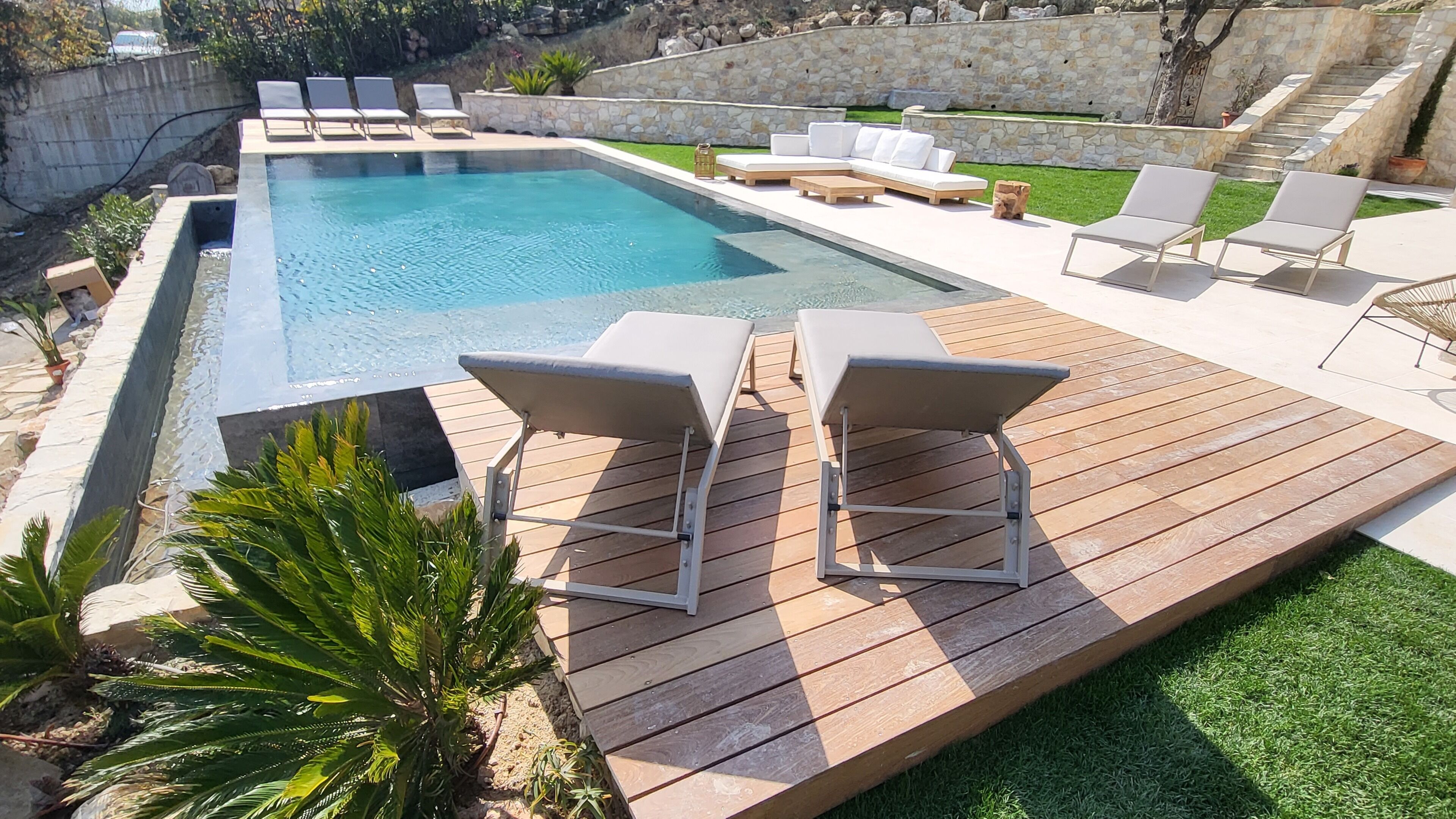 A rectangular swimming pool with a wooden deck, surrounded by lounge chairs and a stone wall.