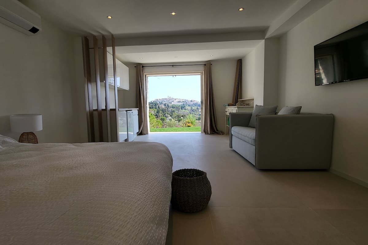 A modern hotel room with a large bed, a sofa, a TV, and a view of a hill.