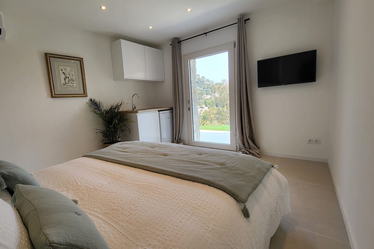 A bedroom with a bed, a TV mounted on the wall, a window with curtains, and a small kitchenette area.