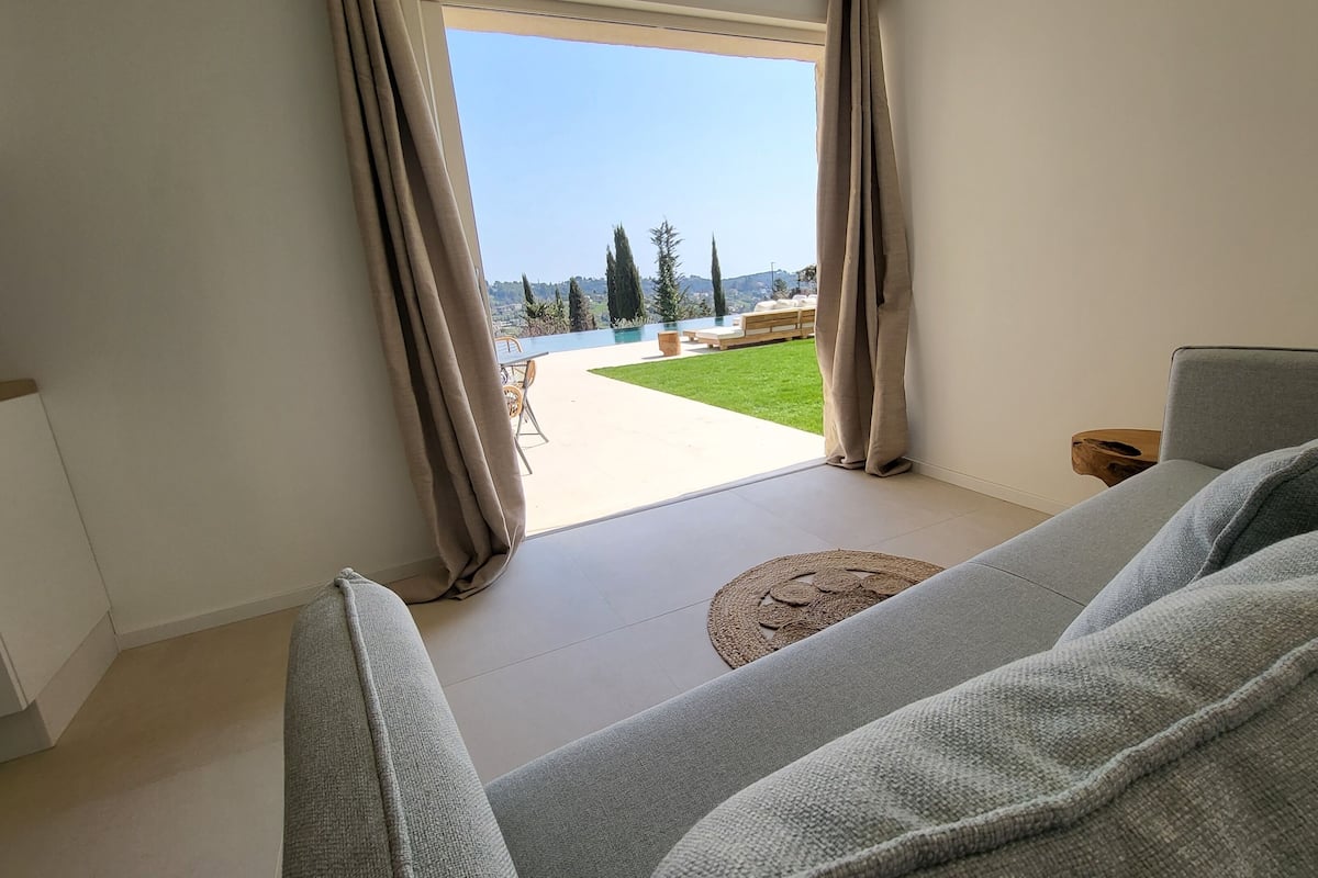 A modern living room with a grey sofa, a small round rug, and a view of a garden and pool through a large window.