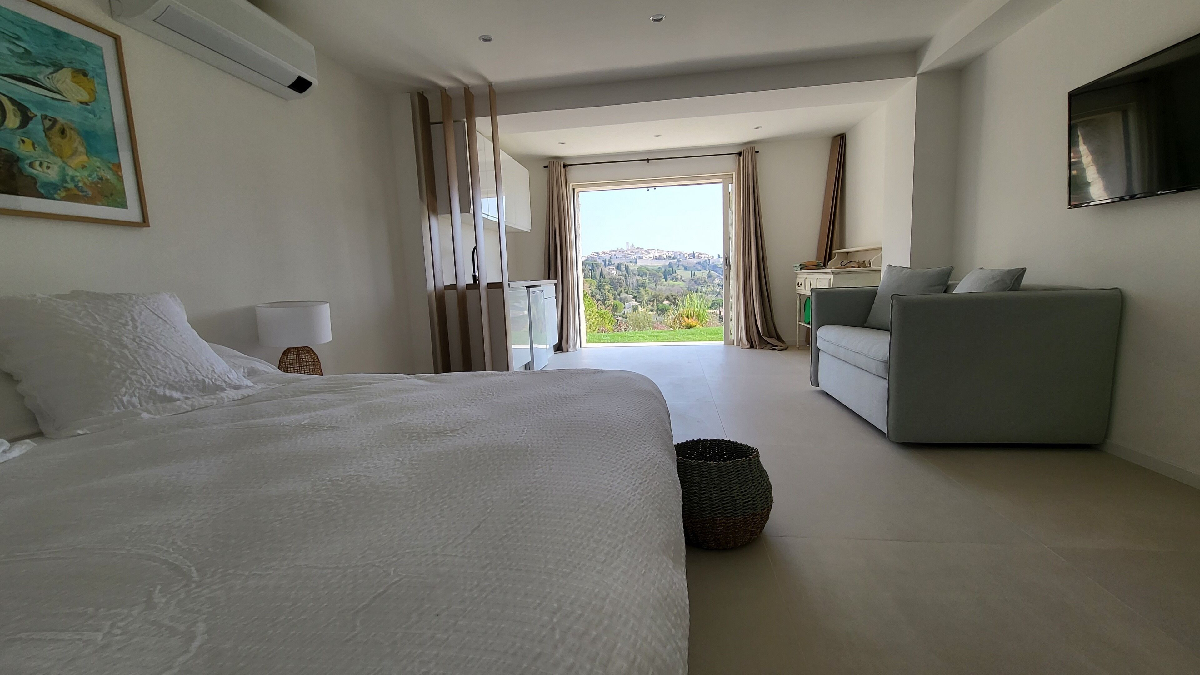 A modern hotel room with a large bed, a sofa, a TV, and a view of a cityscape through the window.