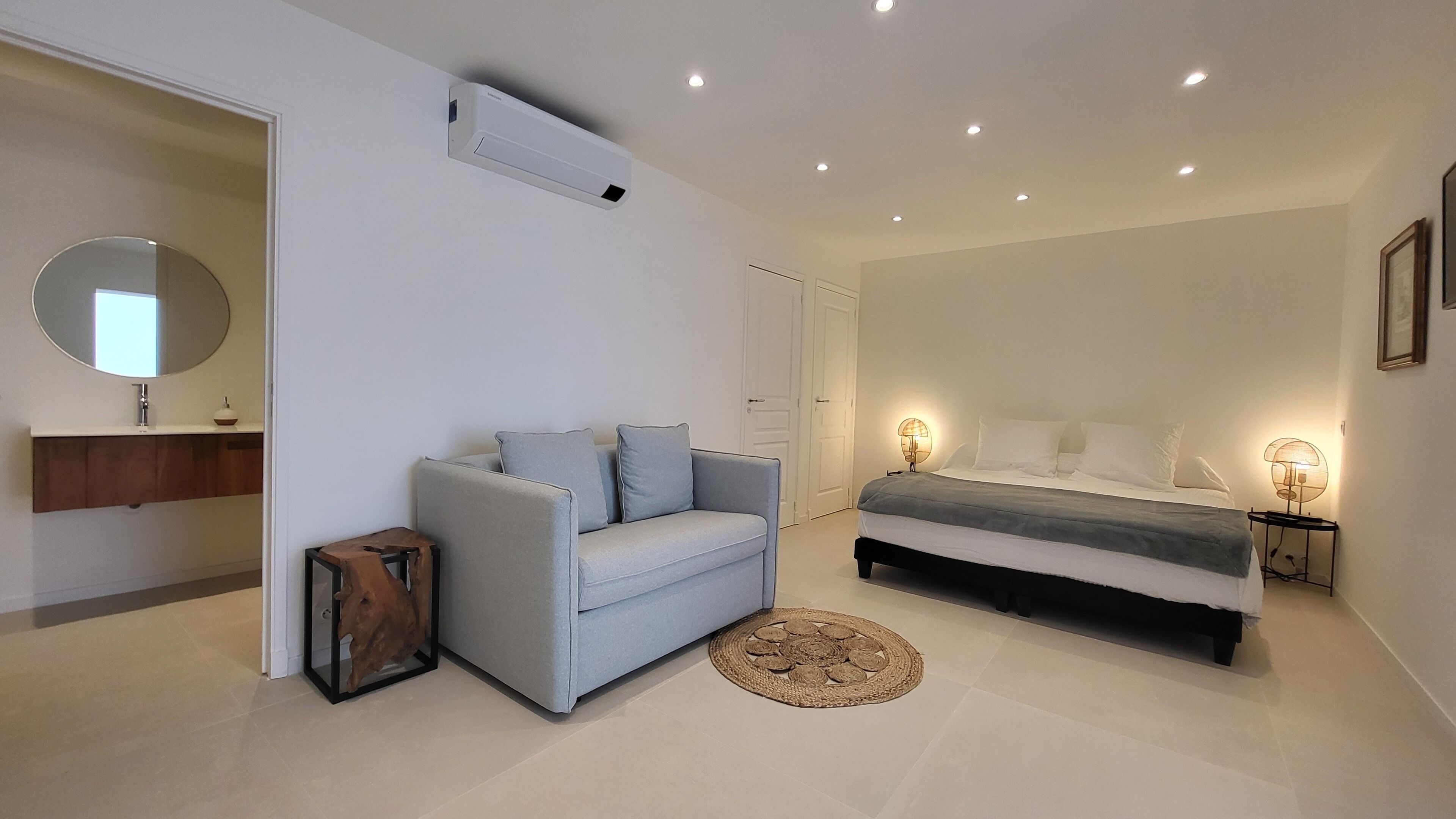 Une chambre à coucher moderne comprenant un lit, un canapé, une table de chevet avec une lampe, un miroir et un petit tabouret en bois.