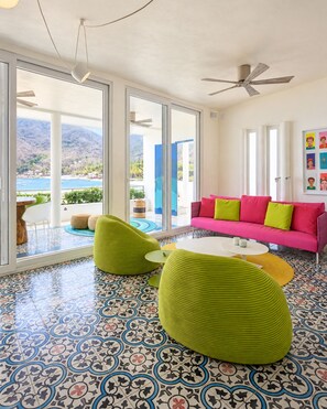 Un salon moderne avec un canapé rose vif, des poufs verts et une grande fenêtre offrant une vue panoramique.