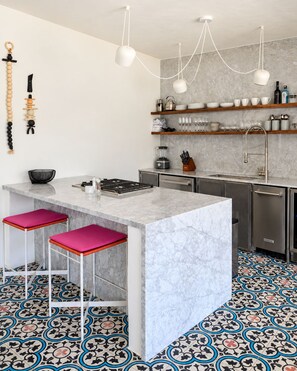 Una cocina moderna con una isla de mármol, dos taburetes de bar, lámparas colgantes y suelo estampado.
