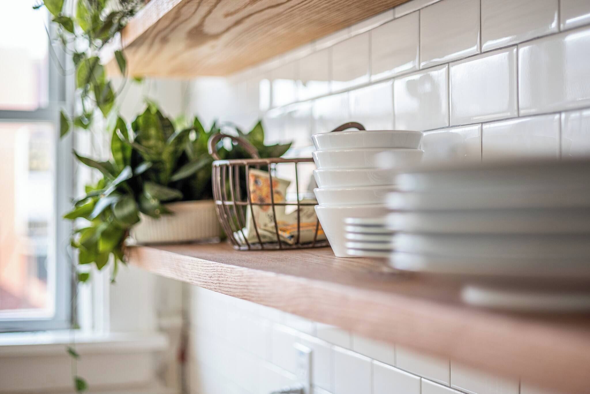 A kitchen counter with white tiles, a wooden shelf, and a plant in a wire basket.