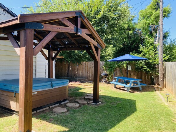 Een houten pergola met een blauwe bank eronder, een picknicktafel en een barbecue in een achtertuin.