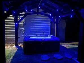 Un jacuzzi bajo una pérgola de madera con luces azules, rodeado de césped y una valla de madera.