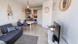 Una sala de estar moderna con un sofá gris, una mesa de comedor con sillas, un televisor y una zona de cocina al fondo.