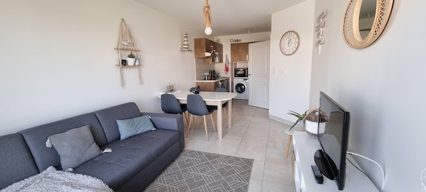 A modern living room with a grey sofa, a dining table with chairs, a TV, and a kitchen area in the background. - Beautiful apartment with sea view 50m from the sea with heated communal pool - Thalasso (Concarneau)