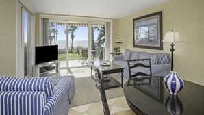 Habitación de hotel con televisor, sofá a rayas, mesa de comedor, lámpara y vista al océano.