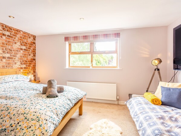 A bedroom with a brick wall, a window with a view, a radiator, a bed with a patterned bedspread, a bedside table with a lamp, and a rolled towel.