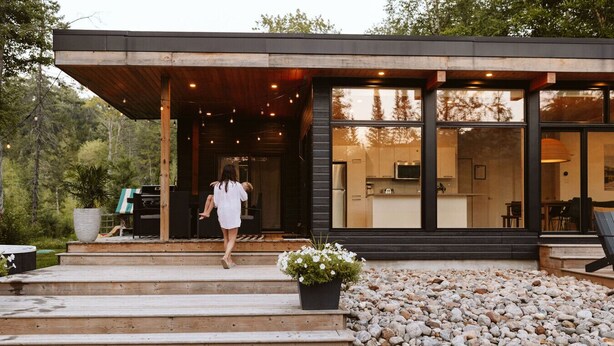 Una casa moderna con un porche cubierto, una cocina visible a través de amplios ventanales y un camino de piedra que conduce a la entrada.