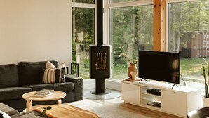 Una sala moderna con una estufa de leña grande de madera negra, un sofá negro, un mueble de televisión blanco y ventanas amplias con vistas al exterior.