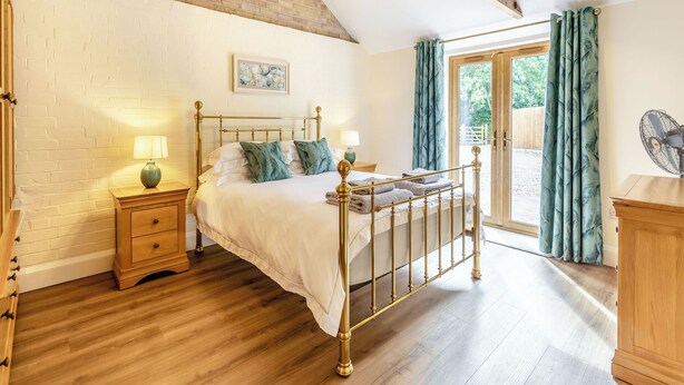 A bedroom with a brick wall, a wooden ceiling, a bed with a metal frame, a bedside table with a lamp, and a view through a glass door. - Tawny Acre Barn - UK38614 (South Walton Highway, Wisbech)