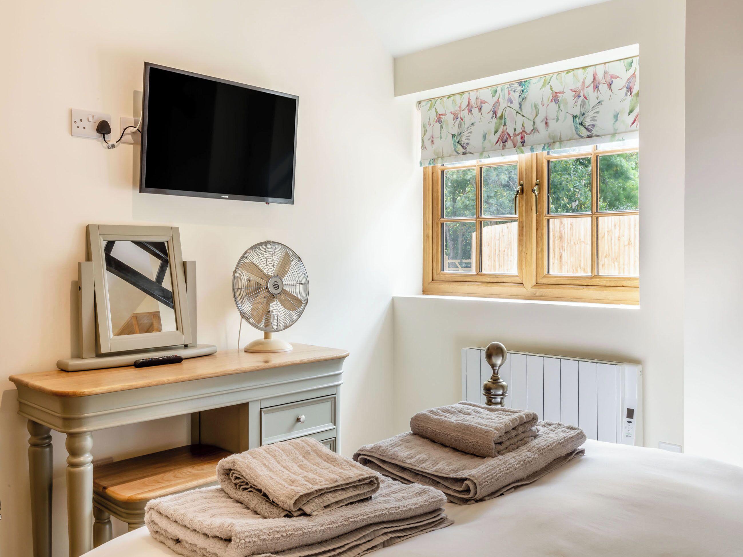 A bedroom with a bed, a bedside table with a TV, a fan, and a window with a view of greenery.