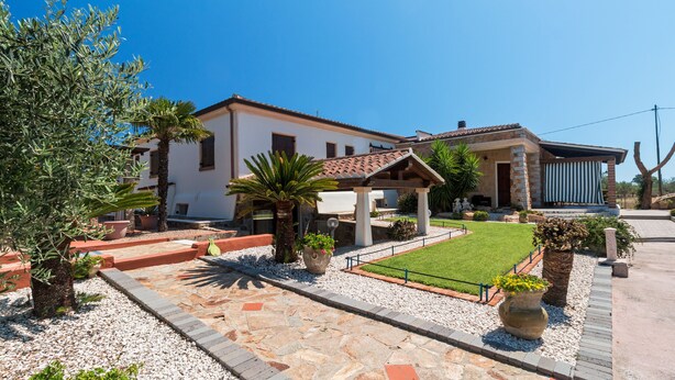 Une villa dotée d’un toit en tuiles, d’un patio couvert et d’un jardin bien entretenu, avec des allées de gravier et des plantes en pot.