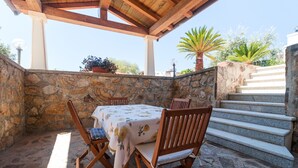 Une terrasse extérieure avec des murs en pierre, une table en bois recouverte d’une nappe à motifs floraux et des chaises en bois.
