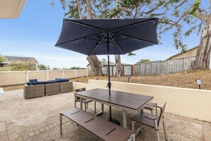 Eine Terrasse im Freien mit Holztisch und -bänken, einem dunklen Sonnenschirm und einer Korbuschlaue mit Kissen.