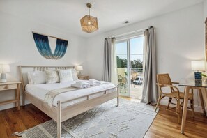 Ein Schlafzimmer mit einem hölzernen Bett, einem Nachttisch, einem Schreibtisch mit Stuhl, einer Pendelleuchte und Blick durch eine Glastür.