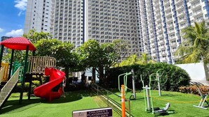 Ein Kinderspielplatz mit einer roten Rutsche, umgeben von grünem Gras und Palmen, mit hohen Wohnhäusern im Hintergrund.