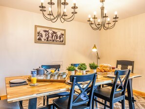 A dining area with a wooden table, chairs, a chandelier, a wall-mounted picture, and breakfast items.