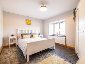 A bedroom with a bed, bedside tables, a window with blinds, a radiator, and a hanging plant.