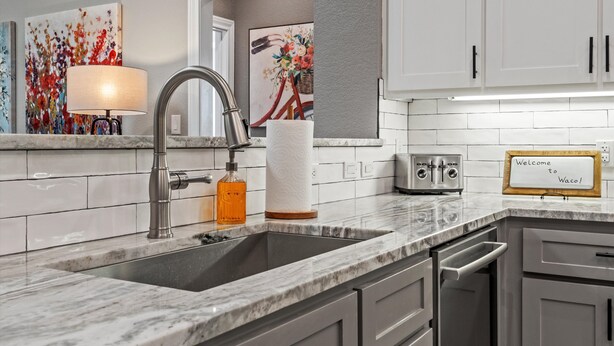 A modern kitchen with a marble countertop, stainless steel sink, and white cabinets. There is a decorative lamp, a framed welcome sign, and a painting on the wall. - Walk to Magnolia • 2 Minutes to Baylor • Huge Private Balcony • King Beds (Waco)
