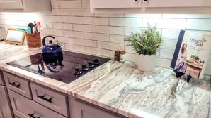 A kitchen with a black induction cooktop, white cabinets, a marble countertop, a potted plant, and a cookbook titled "Magnolia Table". - Walk to Magnolia • 2 Minutes to Baylor • Huge Private Balcony • King Beds (Waco)