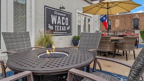 An outdoor patio with a round table, wicker chairs, a fire pit, and a Texas flag. - Walk to Magnolia • 2 Minutes to Baylor • Huge Private Balcony • King Beds (Waco)