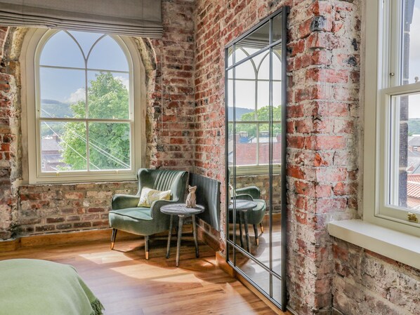 A room with exposed brick walls, large arched windows, a green armchair, a small round table, and a mirror reflecting the room.