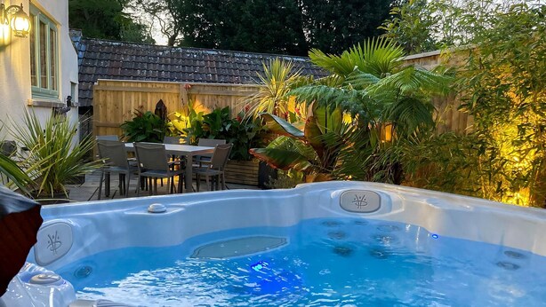 A hot tub in a backyard with a wooden fence, outdoor furniture, and lush greenery.