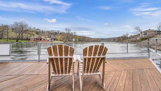 Two wooden Adirondack chairs facing a river with a view of houses and trees in the background.