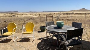 An outdoor seating area with a table and chairs, a large pot, and a view of a dry landscape with distant hills.