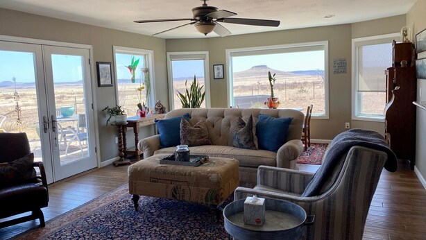 A living room with a sofa, armchair, and ottoman, large windows with a view, and a ceiling fan.