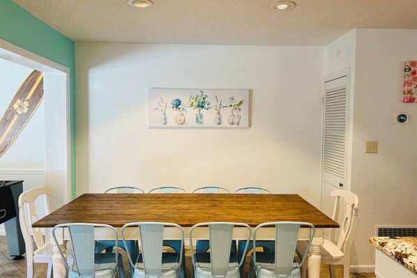Un comedor con una mesa de madera y seis sillas, un cuadro de jarrones con flores en la pared, y una puerta con ventana a la izquierda.