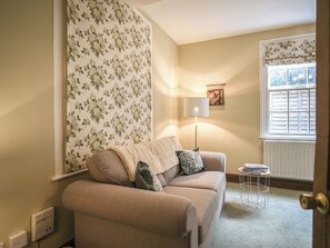 Un salon chaleureux avec un canapé beige, un papier peint à motifs, une lampe sur pied et une fenêtre avec des volets.