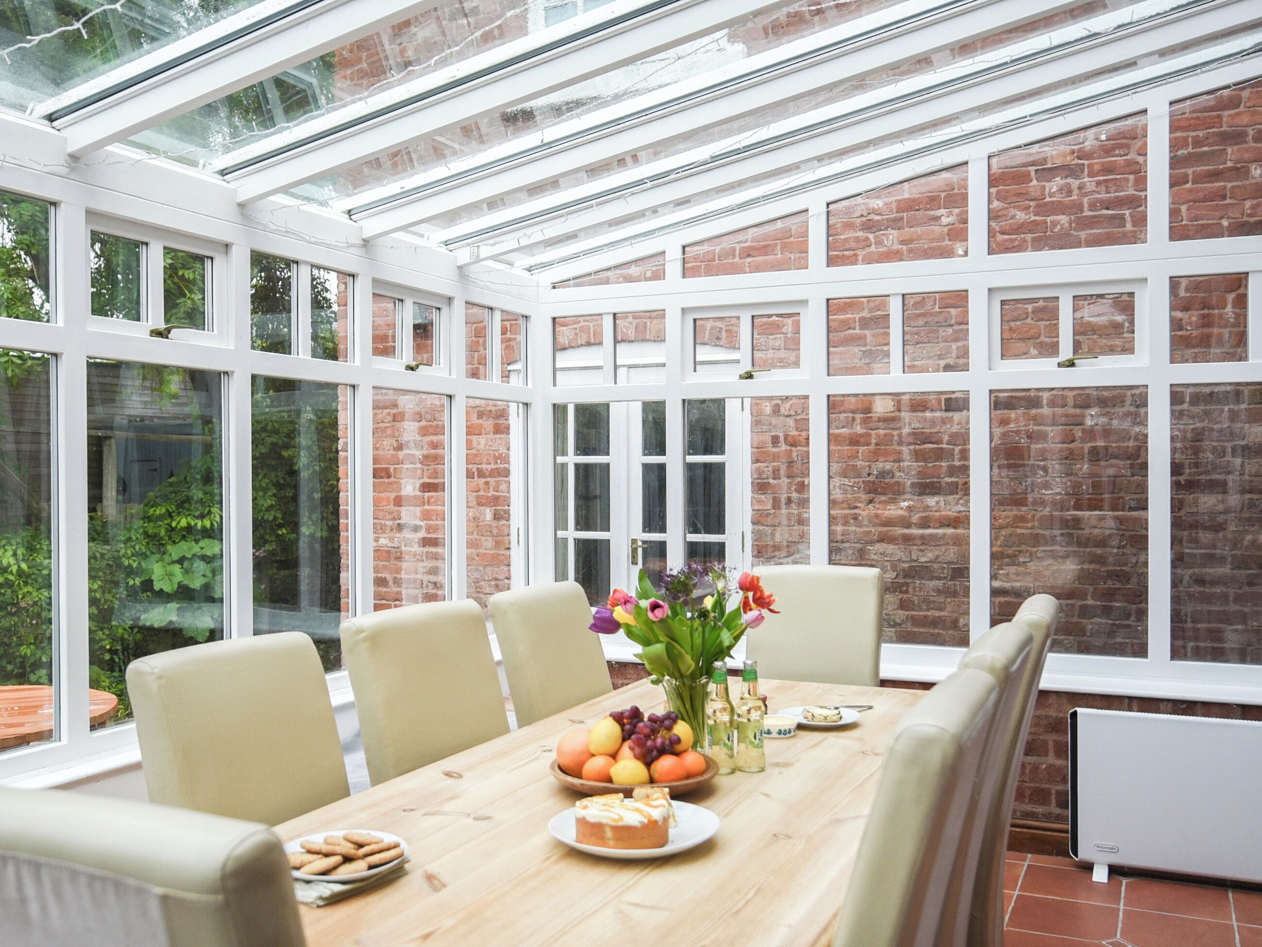 A conservatory with a brick wall and large windows, a wooden table with chairs, and a variety of fruits and flowers.