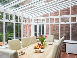 A conservatory with a brick wall and large windows, a wooden table with chairs, and a variety of fruits and flowers.