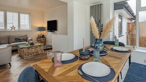A modern living room with a wooden dining table set for two, a sofa, a TV, and a window with blinds. - 3 bedroom accommodation in Rhyl and Prestatyn (Rhyl and Prestatyn)