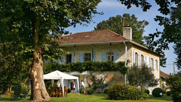A two-story building with a tiled roof, surrounded by greenery and a white tent. - Studio Confort Cerise (Bayonne)