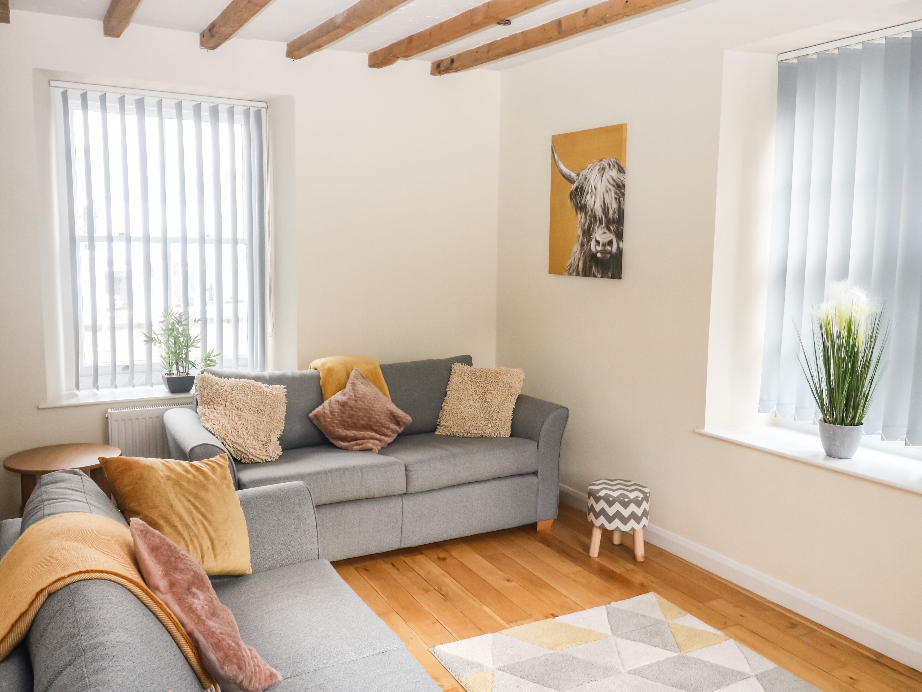 A living room with a grey sofa, wooden beams, a small wooden stool, a potted plant, and a painting of a bull.