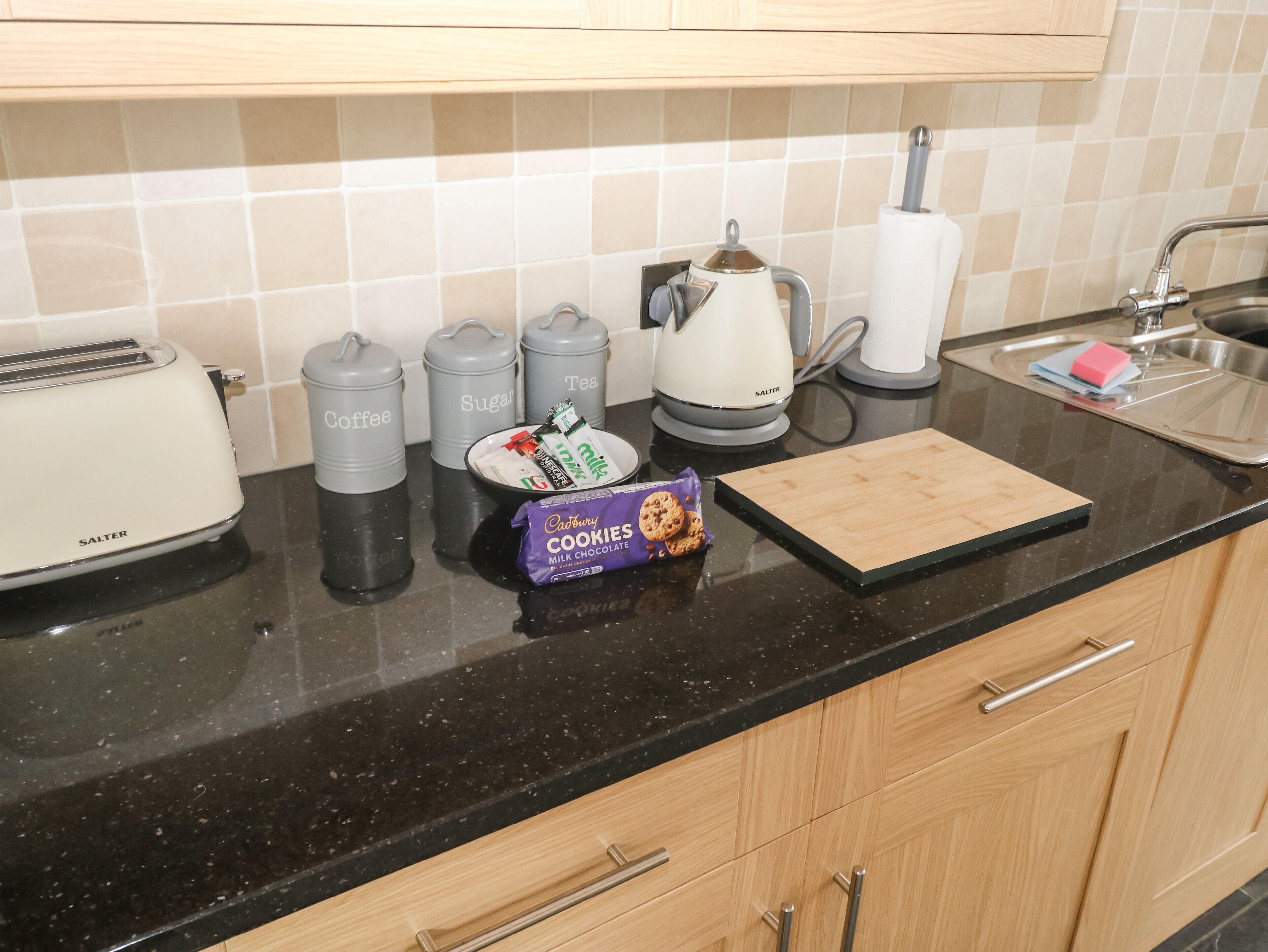 A kitchen counter with a toaster, coffee and sugar containers, a kettle, a plate with cookies, and a cutting board.
