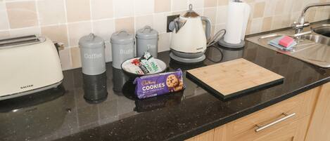 A kitchen counter with a toaster, coffee and sugar containers, a kettle, a plate with cookies, and a cutting board.