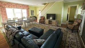 A living room with a sofa, ottoman, and chairs, a television, a staircase, and a dining area.