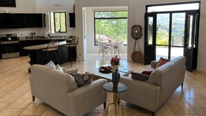 A spacious living area with a kitchen, a dining table, and a sofa. There is a large window providing a view of the outdoors. - Endless charm in the heart of the Central Valley (Atenas)