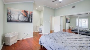 A bedroom with a bed, a ceiling fan, a painting, and a bathroom with a sink and mirror. - The Greenhouse (Houston)